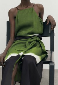 Groene linnen top met dunne bandjes, gecombineerd met een zwarte rok met een witte streep en een strikdetail in de taille. Model zit op een stoel.