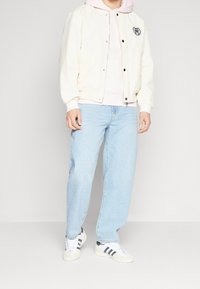 Chaqueta bomber crema clara con un logo redondo, superpuesta a un sudadero rosa pálido. Jeans azul claro y zapatillas blancas con rayas negras.