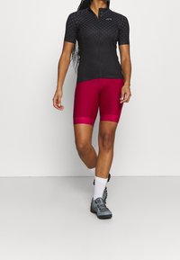 Maillot de ciclismo negro de manga corta con un patrón sutil, combinado con pantalones cortos ajustados de color marrón. Zapatillas de ciclismo en tonos neutros y calcetines blancos.