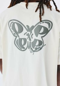 Camiseta color crema con un gran gráfico de mariposa gris en la espalda, acompañado de un texto estilizado que dice "Projects Paris" debajo.