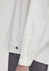 Camicia bianca in lino con una vestibilità rilassata, polsini abbottonati e un dettaglio in bottone nero a contrasto vicino all'orlo. Tessuto morbido e caduta leggera.