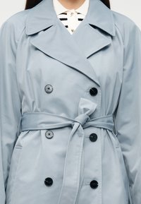 Ανοιχτό μπλε παλτό τύπου trench με διπλό στήθος, μαύρα κουμπιά, μεγάλο γιακά και ζώνη που δένει στη μέση. Μαλακή, λεία υφή υφάσματος.