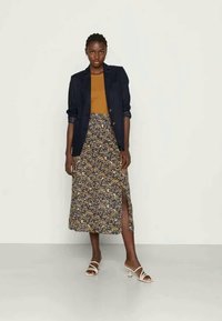 Navy blazer over mustard top, floral midi skirt with side slit. Model wears white strappy sandals. Smooth textures and mixed patterns.