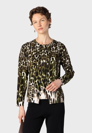 Femme aux cheveux courts et bouclés portant un cardigan à boutons imprimé léopard et un pantalon noir, debout devant un fond uni.