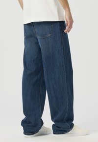 Široke modre kavbojke iz jeansa z zbledele teksture, z dvema zadnjima žepoma in klasičnim krojem, združene z belimi supergami.