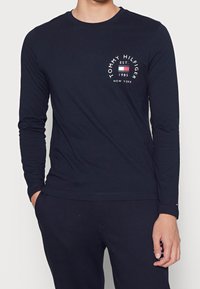 Mies, który ma na sobie czarną koszulę z długim rękawem marki Tommy Hilfiger oraz spodnie z logo i napisem na klatce piersiowej i kieszeni spodni.