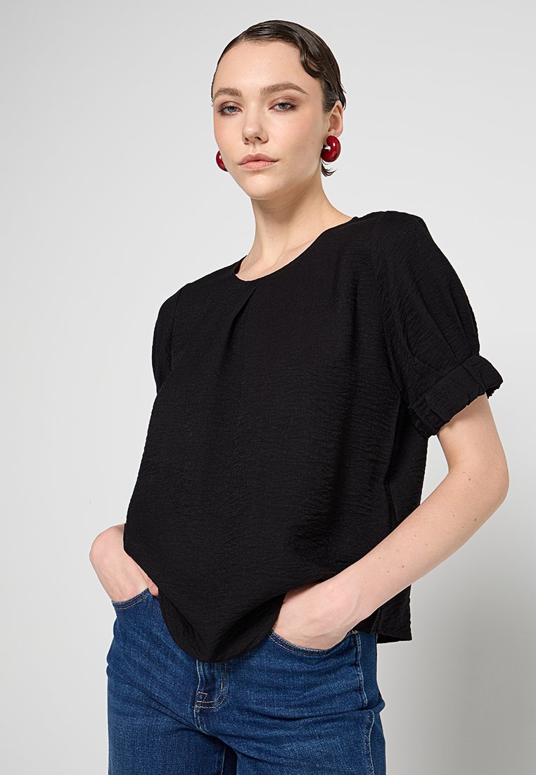Schwarzes, strukturiertes Kurzarmoberteil mit runder Ausschnitt und gerafften Puffärmeln. Kombiniert mit blauen Jeans.