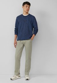 Un giovane in felpa blu a maniche lunghe con motivi di rami, pantaloni beige chiaro e sneaker beige, in piedi contro un muro grigio.