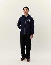 Felpa con zip di colore blu navy, dotata di tasche a marsupio, cappuccio con cordini e logo ricamato sul lato sinistro del petto; abbinata a pantaloni neri e scarpe marroni.