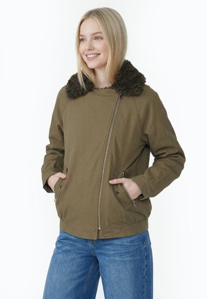Giovane donna con capelli biondi che indossa una giacca verde oliva con colletto in pelliccia nera e jeans blu, sorridente con le mani in tasca.