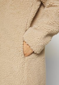 Beiger Fleece-Mantel mit strukturierter Oberfläche, der eine Seitentasche und hochgekrempelte Ärmel für zusätzliche Wärme und Komfort verfügt.
