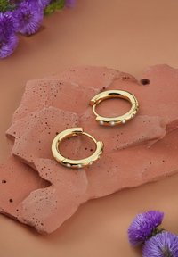 Goldene Creolen mit einem strukturierten Design, die drei kleine Edelsteine besitzen, liegen auf einem terrakottafarbenen Stein Hintergrund mit lila Blumen.