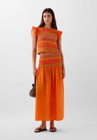 Oranger, taillierter, gesmokter Oberteil mit Rüschensleeves, kombiniert mit einem langen orangefarbenen, gesmokten Rock. Die Accessoires bestehen aus einer braunen Tasche und Sandalen.