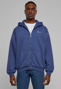 Blå lynlås-hoodie lavet af blødt stof, med en kængurulomme og et lille hvidt logo på venstre bryst. Båret over en hvid t-shirt.