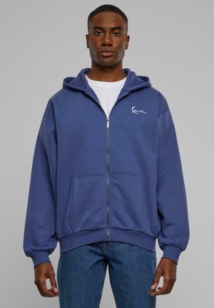 Blue zip-up hoodie made of soft fabric, featuring a kangaroo pocket, and a small white logo on the left chest. Worn over a white t-shirt.
