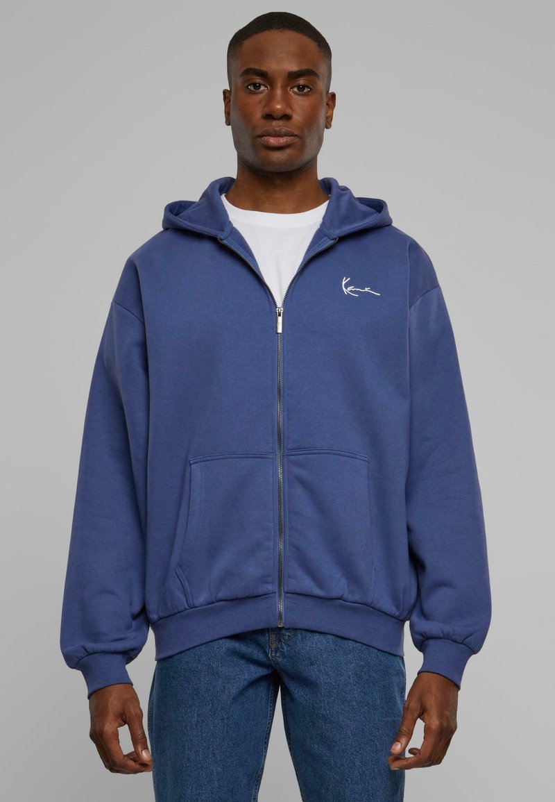 Blå lynlås-hoodie lavet af blødt stof, med en kængurulomme og et lille hvidt logo på venstre bryst. Båret over en hvid t-shirt.