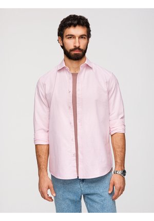 Mand med skæg iført lyserød, åbnet skjorte over en lilla t-shirt og lyseblå jeans mod en ensfarvet baggrund.