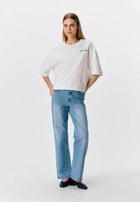 Camiseta corta de manga corta blanca con texto bordado en azul, combinada con vaqueros rectos de color azul claro y zapatos negros sin cordones.