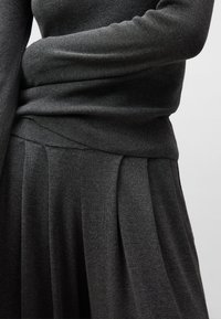 Mörkgrå stickad långärmad tröja i kombination med matchande plisserad kjol, som visar detalj av midjan och vikta armar mot en enkel bakgrund.