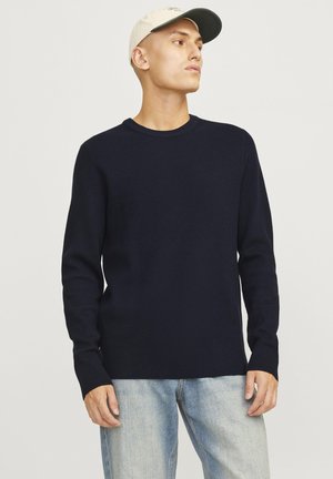 Marineblauer gerippter Pullover mit langen Ärmeln und Rundhalsausschnitt, aus Strickstoff gefertigt. Kombiniert mit hellblauen Jeans und einer beigen Mütze.