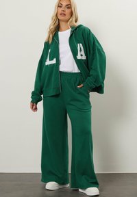 Grüner Kapuzenpullover mit weißer "LA"-Bettung, kombiniert mit passenden weiten Sweatpants, ausgestattet mit elastischem Taillenbund und weißen Sneakers.