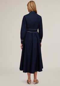 Rochie albastră închisă, cu guler, mâneci lungi și talie ajustată. Prezintă o curea cu detalii de piping și o fustă fluidă.