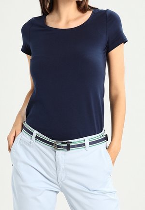 Kobieta ubrana w dopasowany granatowy top z krótkim rękawem, jasnoniebieskie spodnie z paskiem w paski, z rękami luźno w kieszeniach.