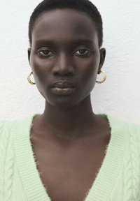 Mäta zelený sveter s vrkočovým vzorom a hlbokým výstrihom do V; model má krátke tmavé vlasy a nosí zlaté kruhové náušnice. Hladká textura pokožky a neutrálny podklad.