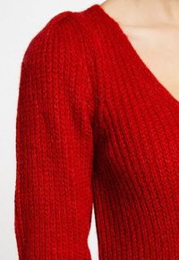 Maglione rosso a maglia con scollatura a V, che presenta un design testurizzato a coste e spalle accentuate. Materiale morbido e caldo.