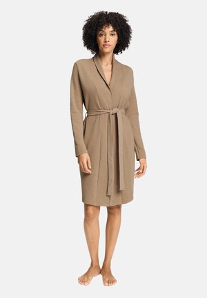 Vrouw met krullend haar, een knielange, beige ochtendjas met lange mouwen, gestrikt in de taille, staand op blote voeten tegen een effen achtergrond.