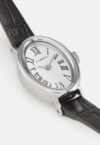 Montre-bracelet en argent avec un cadran blanc, des chiffres romains noirs et un bracelet en cuir noir au fini texturé.