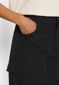 Pantalons cargo noirs avec poches latérales et coutures visibles. Texture douce avec une coupe décontractée. Portés avec une chemise de couleur claire.