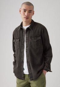 Cămașă din denim gri închis cu un design cu nasturi, două buzunare pe piept și cusături albe, purtată peste un tricou alb și pantaloni verzi.