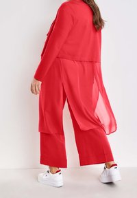 Persona vestida con una larga chaqueta roja y pantalones de pierna ancha a juego, con zapatillas blancas, de pie contra un fondo de pared blanca.