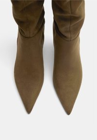 Bottes en daim vert olive, montantes jusqu'au genou, à bout pointu, avec une texture lisse et un design ajusté qui épouse étroitement la jambe.