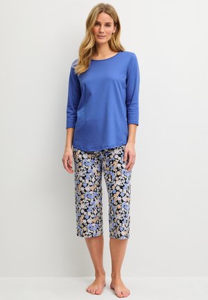 Vrouw staat blootsvoets, draagt een blauwe top met driekwart mouwen en cropped broek met een blauw, wit en bruin bloemmotief.