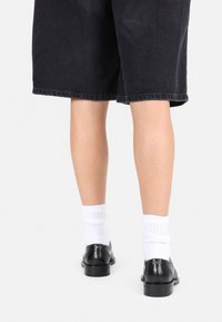 Sorte skinnsko med dekorative detaljer, brukt med hvite anklestrømper. Personen har på seg mørke shorts som viser frem fottøyet.