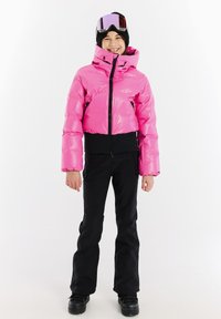 Rosa Pufferjacke mit tailliertem Design, hohem Kragen und schwarzen Akzenten, kombiniert mit schwarzen Skihosen und schwarzen Stiefeln. Schneebrille auf dem Kopf.