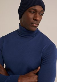 Mornarsko modra pulover s turtleneck iz gladkega pletenega materiala, z visokim ovratnikom in prilegajočim se krojem, nosi zašito pleteno kapo v istem odtenku.