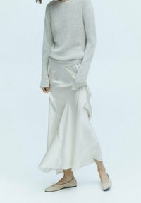 Femme portant un pull en maille gris clair, une jupe midi en satin crème et des ballerines beiges sur un fond blanc uni.
