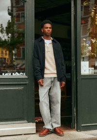 Casual outfit featuring a dark button-up jacket, beige knitted sweater, light gray jeans, and brown lace-up shoes, standing by a green door.