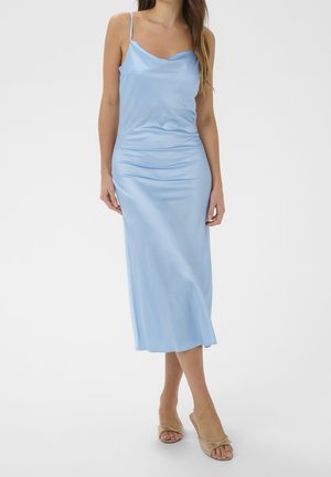 Mujer que lleva un vestido de satén azul claro con tirantes finos, de pie con sandalias de tacón abiertas beige sobre un fondo blanco liso.