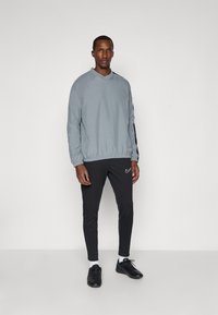 Lys grå pullover med høj krave og elastisk kant, parret med sorte slim-fit bukser med et lille logo. Sorte træningssko.