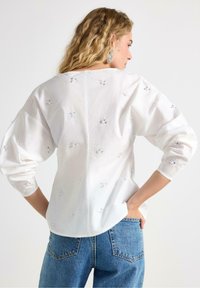 Vit långärmad blus med broderade blommönster och silverdetaljer, kombinerad med blå högmidjade jeans i denim. Slät tygstruktur.