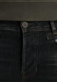 Jeans de mezclilla oscura con una sutil textura, costuras naranjas en contraste y un botón de metal en la cinturilla. Cintura verde acanalada visible por encima.