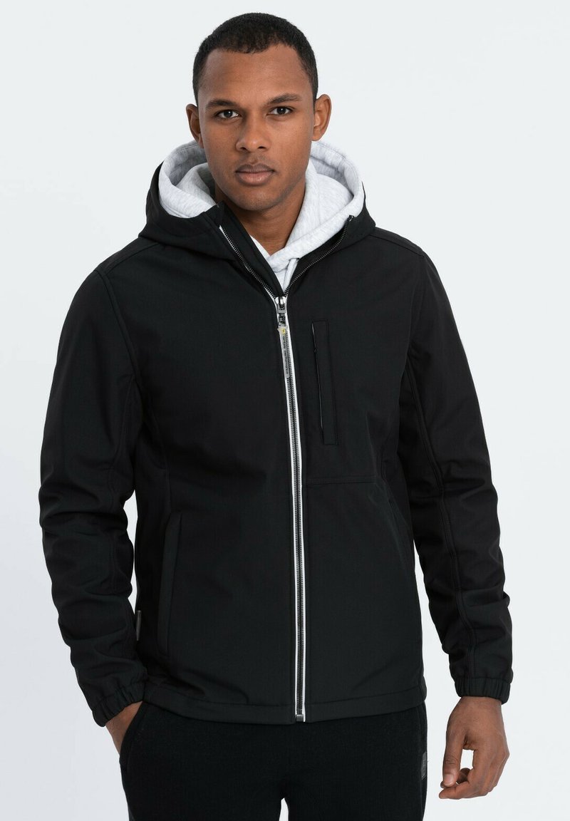 Veste à capuche noire avec une fermeture éclair à l'avant, dotée d'une poche poitrine et d'une doublure intérieure grise. Tissu lisse et poignets élastiques.