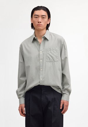 Młody mężczyzna z zaczesanymi do tyłu czarnymi włosami, ubrany w jasnoszarą koszulę na guziki i czarne spodnie, stojący na tle jednolitej białej ściany.