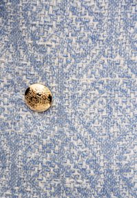 Jasnoniebieska i biała tekstura materiału z wzorem tkanym, z złotym guzikiem o skomplikowanym wzorze na środku.