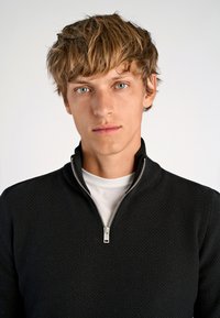 Maglione zip-up nero con texture, colletto alto, indossato sopra una camicia bianca, con dettagli in cerniera argentata. Lo sfondo è semplice.