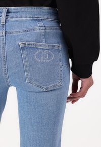Jeans de denim azul claro con un diseño ajustado, que cuentan con un bolsillo trasero marcado por un logo decorativo hecho de pequeños studs.